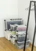 A stack of clear plastic storage boxes with black lids and handles, some containing folded clothes and other items, are placed in a corner against a white wall. A black metal coat rack with hangers is visible on the right side of the image. Produktbild-Vorschau