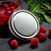 A close-up shot of a glass jar filled with raspberries, with its silver lid placed next to it. Several fresh raspberries and green leaves are scattered around the jar on a dark, textured surface. Produktbild-Vorschau