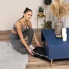 A woman in athletic wear is kneeling on a rug next to a blue coffee table, placing a black item into an open compartment of the table. Produktbild-Vorschau
