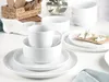 A collection of white ceramic dishes, including bowls, plates, and a mug, arranged on a white surface. Some crackers with toppings are visible on a plate in the background. Produktbild-Vorschau
