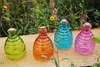 Four colorful glass bottles with cork stoppers and wire handles are arranged on a wooden surface in front of a blurred background of green foliage and colorful flowers. Produktbild-Vorschau