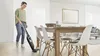 A woman is vacuuming a dining room floor with a cordless stick vacuum cleaner. Produktbild-Vorschau