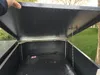A person's hand is opening the lid of a dark gray trash bin enclosure, revealing a black trash can inside. The enclosure is outdoors, with a grassy area and trees visible in the background. Produktbild-Vorschau