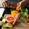 A close-up shot of hands juicing an orange with a wooden juicer, surrounded by fresh citrus fruits and a glass of orange juice. Produktbild-Vorschau