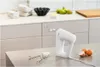 A white hand mixer with its attachments is placed on a metallic surface. In the background, there are ingredients for baking on a kitchen counter. Produktbild-Vorschau