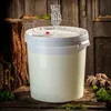 A white fermentation bucket with a clear airlock and stopper sits on a rustic wooden surface, surrounded by natural elements like wood and moss. Voir les images du produit