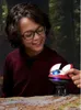 A young boy wearing glasses and a maroon hoodie is looking at a small, red and white electronic toy droid with a blue light on its head. The toy is sitting on a surface that looks like dirt and rocks, with a blurred forest background. Anteprima dell’immagine prodotto