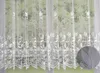 A white lace curtain with a floral pattern is shown hanging in front of a window with greenery visible outside. Produktbild-Vorschau