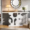 A white console table with a decorative base is shown in a well-lit room, with a lamp, framed pictures, and a vase of flowers on top. A large round mirror hangs on the wall behind it. Anteprima dell’immagine prodotto