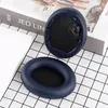 Two blue ear pads for headphones are placed on a black and white checkered surface, with a book in the background. Produktbild-Vorschau