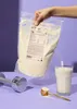 A hand holding a bag of vegan protein powder next to a glass of protein shake, a scoop, and a dumbbell. Produktbild-Vorschau