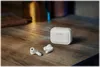 White wireless earbuds and their charging case are placed on a wooden surface, with soft lighting in the background. Anteprima dell’immagine prodotto