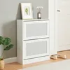 A white shoe cabinet with two flip-down doors, placed in a room next to a wall and a doorway. A framed picture and a vase with flowers are on top of the cabinet. A plant is to the left, and a pair of shoes is on the floor to the right. Podgląd zdjęcia produktu