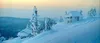 A snowy landscape with two small houses covered in snow, with snow-covered trees in the foreground. Náhľad obrázku produktu