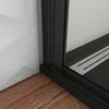 A close-up view of the bottom corner of a shower enclosure, showing the black frame, a wooden floor, and a white wall. Produktbild-Vorschau