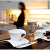 A person is in the background of a kitchen, while coffee cups and a glass are in the foreground. Produktbild-Vorschau