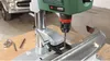 A drill press is shown drilling into a metal object, possibly a car bumper, on a wooden surface. Other drill bits and a metal piece are visible on the surface. Podgląd zdjęcia produktu