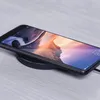 A smartphone is placed on a black wireless charging pad on a light-colored wooden surface. Produktbild-Vorschau