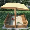 A wooden sandbox with a canopy, benches, and some toys, set in a grassy backyard with trees in the background. Podgląd zdjęcia produktu