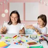 Two young girls are sitting at a table, engaged in a craft activity with various art supplies spread out before them. Produktbild-Vorschau
