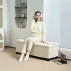 A young woman is sitting on a beige storage bench in a well-lit room, wearing headphones and listening to music. Podgląd zdjęcia produktu