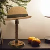 A hat stand with a hat on it, placed on a dark surface with some greenery and oranges in the background. Produktbild-Vorschau