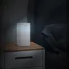 A white aroma diffuser with a blue light sits on a wooden nightstand next to a bed. Produktbild-Vorschau