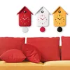 Three cuckoo clocks in different colors (red, white, yellow) are displayed on a white wall above a yellow couch with red cushions. Produktbild-Vorschau