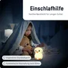 A young girl is under a blanket with a teddy bear, illuminated by a small, glowing duck-shaped night light. Produktbild-Vorschau