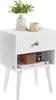 A white nightstand with a drawer and an open shelf, decorated with a vase of greenery, a cup of coffee, and a small radio on a book. Produktbild-Vorschau