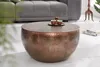 A round copper coffee table with hammered detailing is shown in a living room setting, with a sofa in the background and a small set of coffee cups on top of the table. Produktbild-Vorschau