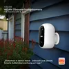A white security camera mounted on a blue, clapboard-sided house, with red-leafed plants in the foreground. Produktbild-Vorschau