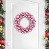 A pink Christmas ball wreath is hanging on a white door, with a decorated Christmas tree visible on the left side. Voir les images du produit
