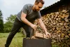 A man is placing firewood into a felt bag next to a large stack of firewood and a wooden building. Náhled obrázku produktu