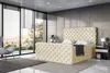 A modern bedroom with a large window overlooking a scenic view, featuring a tufted beige boxspring bed with a mattress and topper, a gray armchair, and a bedside table. Produktbild-Vorschau