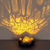 A solar-powered lantern with a cracked glass orb inside a lotus-shaped metal base, casting intricate shadows on a wall. Produktbild-Vorschau