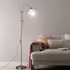 A floor lamp is positioned next to a sofa with pillows and a blanket. Produktbild-Vorschau