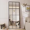 A full-length mirror with a black grid frame is placed in a room, reflecting a sofa, a lamp, and a painting on the wall. Produktbild-Vorschau