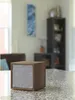 A wooden cube speaker with a gray fabric front sits on a speckled countertop in a living room setting. Produktbild-Vorschau