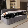 A dark gray upholstered bed frame with the mattress lifted to reveal storage space underneath, containing folded clothes and a bag. The bed is in a bedroom setting with shelves on the wall behind it. Anteprima dell’immagine prodotto