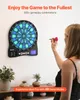 A woman is about to throw a dart at an electronic dartboard mounted on a wall. The dartboard has LED lighting and displays game information. Decorative plants and vases are on a shelf below. Produktbild-Vorschau