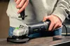 A person is holding an angle grinder, preparing to attach the auxiliary handle. Náhled obrázku produktu