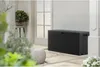 A dark grey outdoor storage box is placed on a patio next to a white door. A potted plant is visible on a stool to the left, and a textured ottoman is in the foreground. Náhľad obrázku produktu