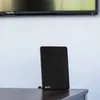 A black Vayox DVB-T/T2 antenna is placed on a dark wooden surface in front of a white wall and a blurred television screen. Podgląd zdjęcia produktu