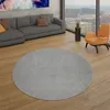 A modern living room with a grey sectional sofa, an orange accent chair, and a large circular grey rug on a wooden floor, with a cityscape visible through a large window. Produktbild-Vorschau