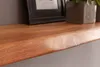 A close-up view of a wooden shelf with a natural edge, showing the wood grain. A framed picture and a potted plant are visible in the background. Náhled obrázku produktu