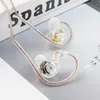 A pair of transparent in-ear monitor headphones with gold and silver accents, resting on a surface with a book in the background. Produktbild-Vorschau