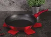 A red frying pan with a black speckled interior sits on a dark surface, supported by a red trivet. A potted plant is visible in the background. Náhled obrázku produktu