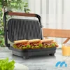 A sandwich maker with three sandwiches inside, placed on a table with a glass of orange juice and some lettuce in the foreground. Produktbild-Vorschau