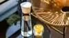 A glass carafe filled with water and a glass with lemon slices, placed on a glass table next to a wicker chair. Produktbild-Vorschau